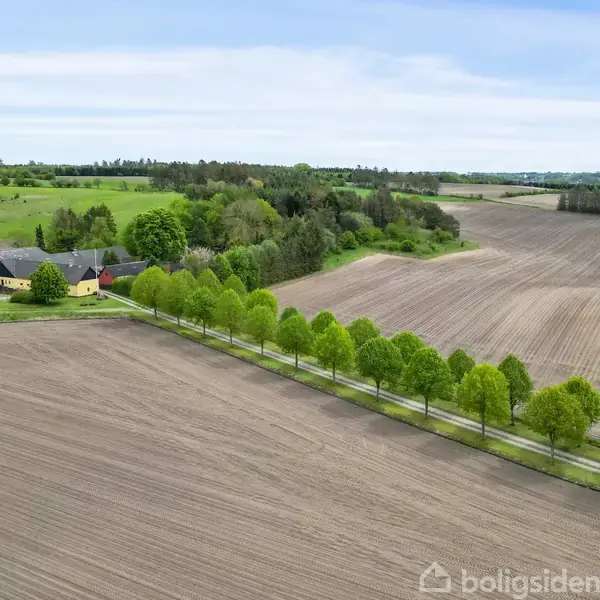 En gård ligger stille mellem grønne træer ved siden af en nyligt pløjet mark. Omkring gården er der områder med skov og marker, der strækker sig til horisonten.