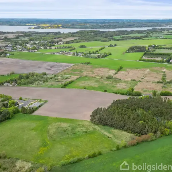 Landområde markeret af marker i forskellige nuancer af grønt og brunt strækker sig ud i landskabet. En sø og flere skovområder findes i baggrunden under en overskyet himmel.