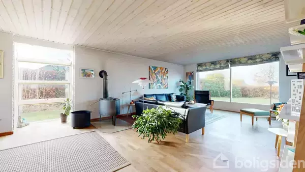 Stue med sofagruppe i hjørnet, pejs til venstre, samt store vinduer, der giver udsigt til have. Rummet har lyst trægulv og hvide trævægge og loft, dekoreret med kunst.