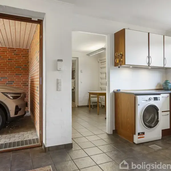 En vaskemaskine står i et bryggers med hvide skabe og gråt klinkegulv. Til venstre en åben dør ind til en garage, hvor en bil er parkeret.