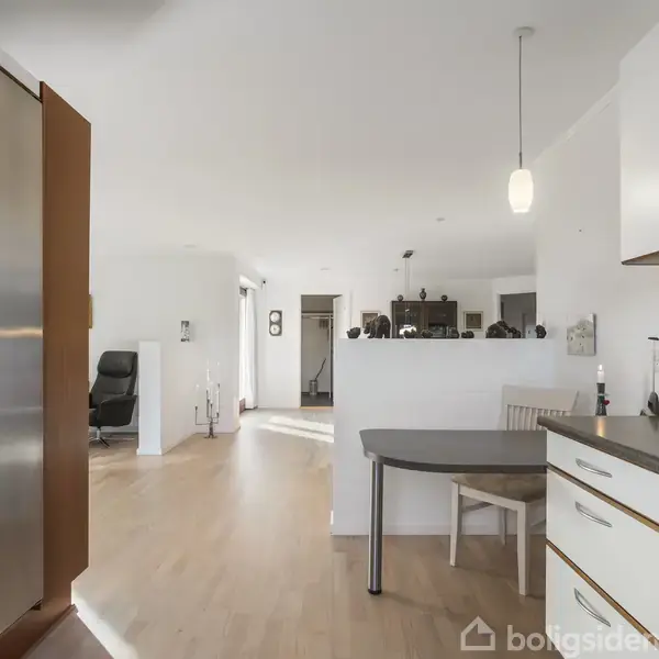 Køkken med hvide skabe og mørk bordplade. Halvø med indbygget bord og stole. Lys trængt fra pendellampe. Åben forbindelse til stue, trægulv og moderne møblering.