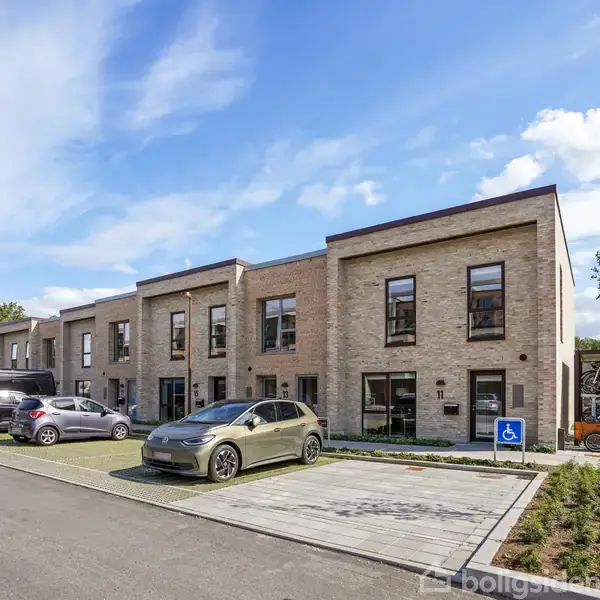 Rækkehuse i moderne stil med fladt tag, stående langs en parkeringsplads med biler, herunder en handicapparkeringsplads. Beliggende i et grønt område under en blå himmel.