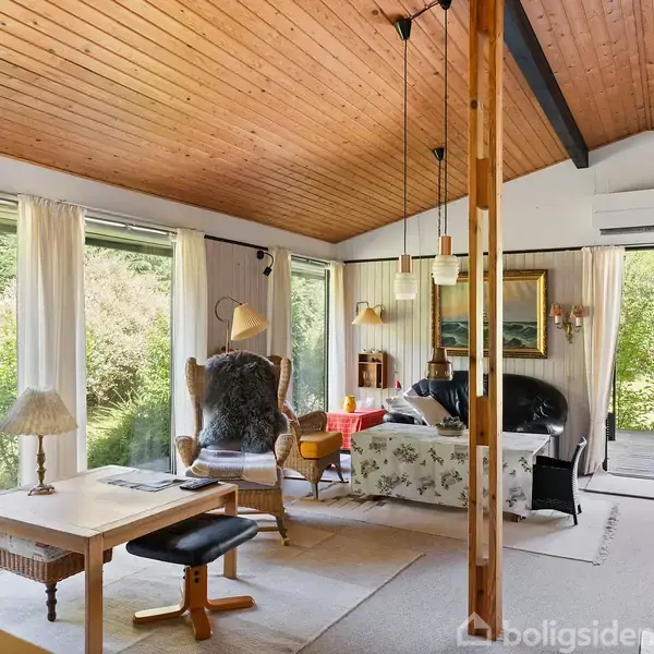 Stue med træloft, store vinduer og sofaområde. Spisebord med stole til venstre. Rummet har udsigt over en grøn have gennem vinduerne.