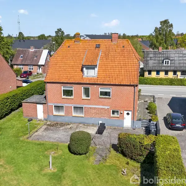 Rødt murstenshus med rødt tegltag står stille i et parcelhuskvarter med velplejede haver og parkerede biler. Surroundet af hække, og der er flere omkringliggende huse i baggrunden.
