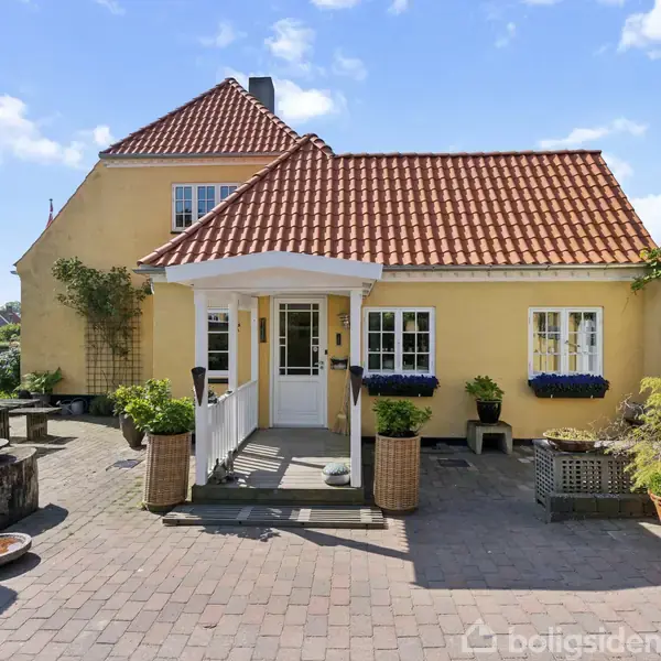 Gult hus med rødt tegltag står i solskin. Foran indgangen er potteplanter og blomsterkasser, omgivet af en stenbelagt gårdsplads med siddeområde. Blå himmel og naboer i baggrunden.