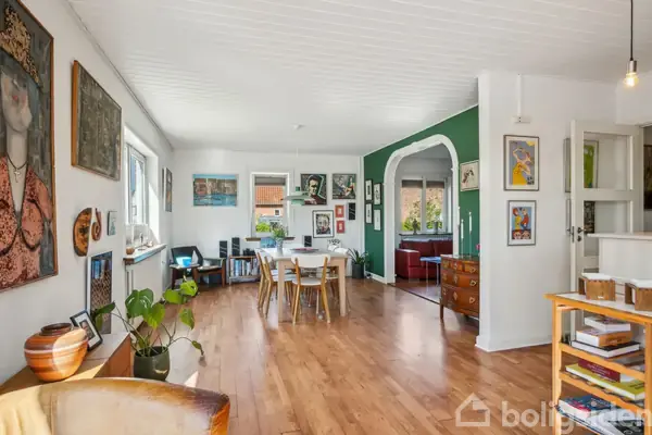 Spisestue med træspisebord og stole står i centrum, omringet af kunst på væggene. Åben bue fører til stue med sofa. Lys strømmer ind fra vinduer i moderne, farverigt hjem.