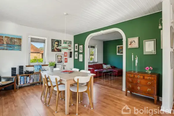 En spisestue med et træbord og seks hvide stole. I baggrunden ses en stue med en rød sofa. Væggene er dekoreret med forskellige plakater og malerier.