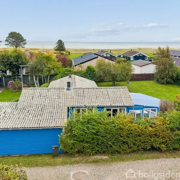 Blåt hus står stille i grønne omgivelser med hække og buske. I baggrunden ses flere huse og en kystlinje under en klar, blå himmel.