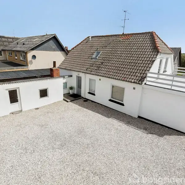 Et hvidt hus med rødt tegltag står i en grusbelagt gård. Der er en stor terrasse ovenpå en tilbygning. Omkransende bygninger ses i baggrunden.