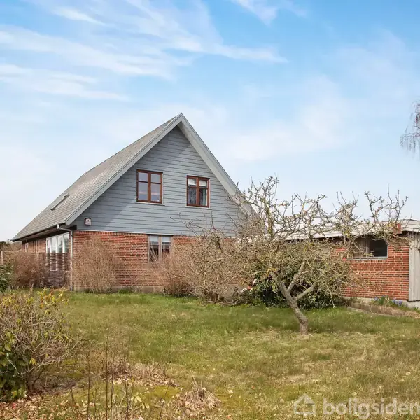 Et to-etagers hus med trekantet tag står stille omkranset af en have med træer og buske. Skuret til højre og blå himmel danner baggrunden.