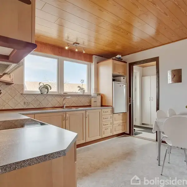 Køkken med lyse træskabe og grå bordplader, udstyret med en komfur. To store vinduer lader lys ind. Ved siden af er et spisebord med hvide stole og en blomsterdekoration.