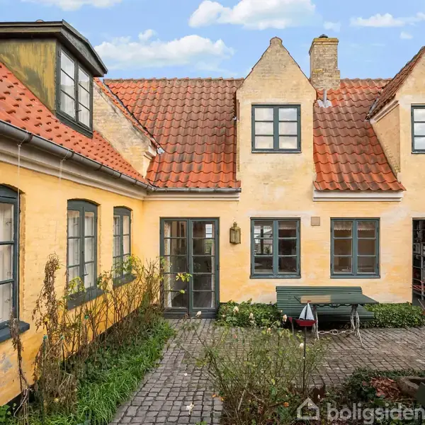 Gult, charmerende hus med rødt tegltag, to kviste og flere vinduer. Står rundt en brostensbelagt gårdhave med grønt buskads og havemøbler. Klar himmel ses i baggrunden.