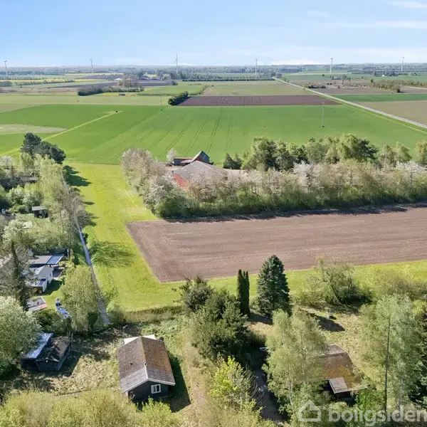 Et landligt område med marker, hvor en gård er omkranset af træer. I forgrunden ses små bygninger blandt frodige træer. Vindmøller står i horisonten under en klar himmel.