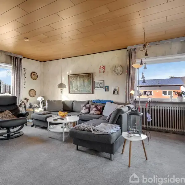Stue med gråt hjørnesofaarrangement fyldt med puder, sofabord med bøger, og sort lænestol ved siden af. Store vinduer giver udsigt til et boligområde udenfor.