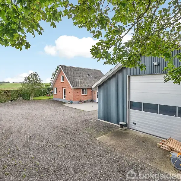 Rødt murstenshus står stille ved siden af en stor blå garage med rulleport, omgivet af grusvej og grønne marker, set under skyggen af et træ med grønne blade.