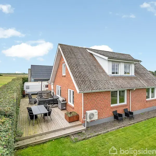 Rødt murstenshus med skråtag har en træterrasse med havemøbler. Huset ligger på en græsplæne, omgivet af hække, med åbne marker i baggrunden under en blå himmel.