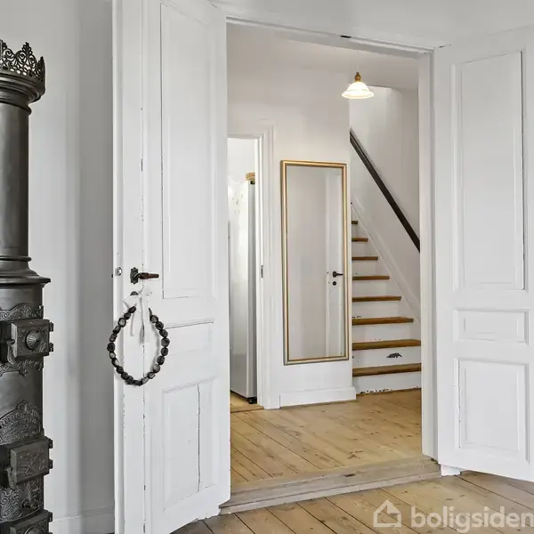 Brændeovn står stille ved en åbning mellem to rum med hvide døre. Trægulv går mod en trappe oplyst af en loftlampe, der fører til en anden etage.