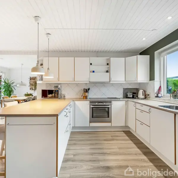 Et lyst køkken med hvide skabe og trægulv. Der er en stor ø med barstole og pendellamper. Stor vindue giver udsigt til en grøn have.