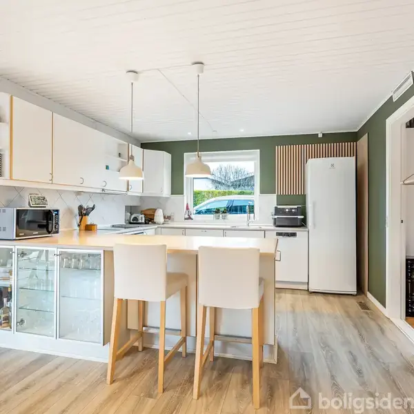 Køkkenø med to barstole og pendellamper, omgivet af køkkenskabe og hvidevarer. Rummet har grønne vægge og trægulv, med dekorationer og køkkenredskaber på hylder og borde.