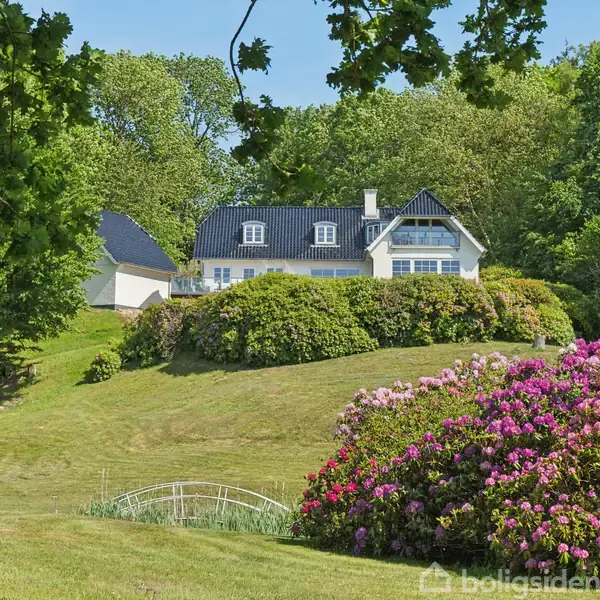 Et hvidt hus med sort tag står i en frodig have med farverige blomster og buske. En lille bro krydser en lavning i græsset, omgivet af træer i baggrunden.
