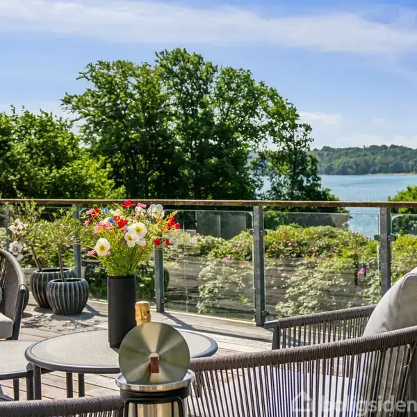 Terrassemøbler er stillet på en træterrasse med udsigt over en sø. En vase med blomster står på et bord, omgivet af frodigt grønt og træer i baggrunden.