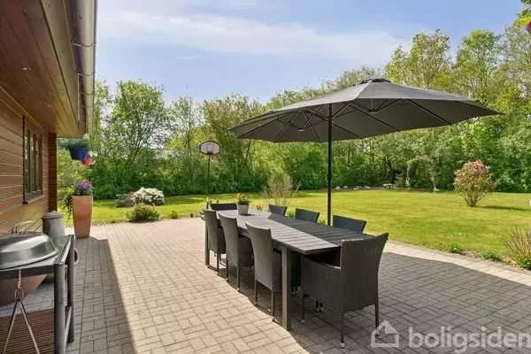 Terrassemøbler med sort parasol står på en flisebelagt terrasse. I baggrunden ses en stor, grøn have omgivet af træer. En grill er placeret ved husvæggen til venstre.