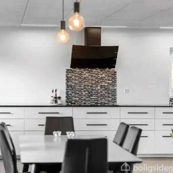 Et moderne køkken med hvide skabe. Sorte stole står ved et bord. Baggrunden har en sort emhætte over en flisebelagt væg med mosaikmønster. Tre pendellamper hænger fra loftet.