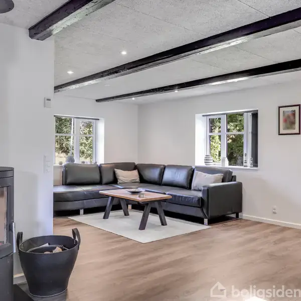 Brændeovn med levende ild står i et moderne stueområde med sort lædersofa, træbord og trægulv. To vinduer giver naturligt lys; væggen har kunstværk og en kommode.