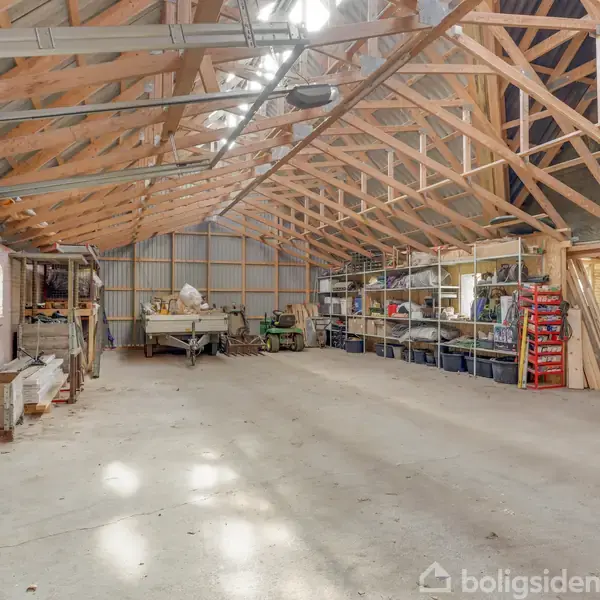 En rummelig garage med træloftsbjælker og betonbund, indeholder redskaber og materialer på hylder langs væggen, samt en garageport til venstre. Rummet er lyst og åbent.