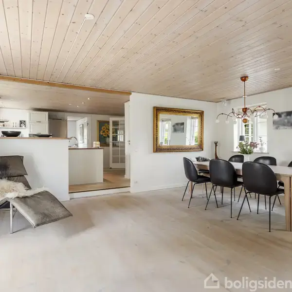 Spisebord med seks stole står i en lys stue med træloft og lyse trægulve. En grøn plante, spejl og køkken i baggrunden giver en indbydende atmosfære.