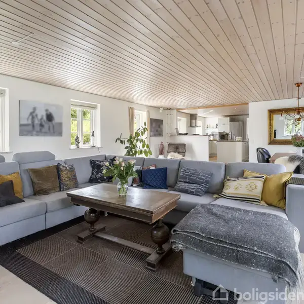 Stue med en stor, lys hjørnesofa fyldt med farverige puder. Et træbord står foran sofaen. I baggrunden ses et åbent køkken og en spisestue med loftslampe.