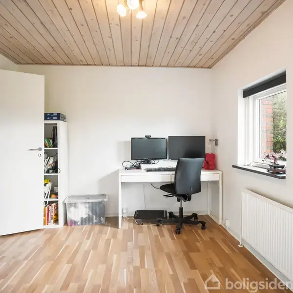 En skrivebordsstol står foran et skrivebord med to skærme og en computer i et rum med parketgulv og et træloft. Hylder og en åben dør ses i baggrunden.