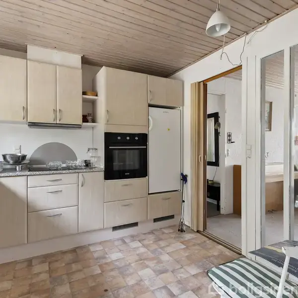 Køkken med lyse træskabslåger og indbygget ovn ved siden af køleskab. Der er porcelæn og køkkenredskaber på bordpladerne. Rummet har lyse fliser og et trægulv.