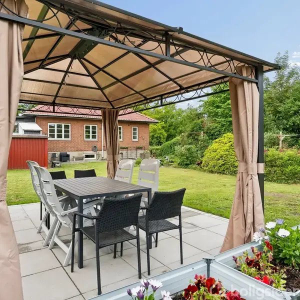 Pavillon med spisebord og stole står på flisebelagt terrasse; omgivet af blomsterkasser og græsplæne. En rødbygget murstensbygning ses i baggrunden, omgivet af grønne træer og buske.