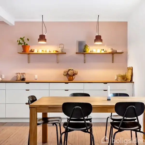 Et spisebord med fire sorte stole står i et moderne køkken. Over bordet er to hylder med dekorative genstande og planter oplyst af loftlamper. Væggene er lyserøde.