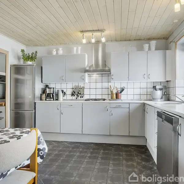 Et moderne køkken med grå skabe og apparater udfører madlavning. Rummet har hvide fliser, et spisebord med blomstret dug og stole, og det er oplyst af loftlamper.