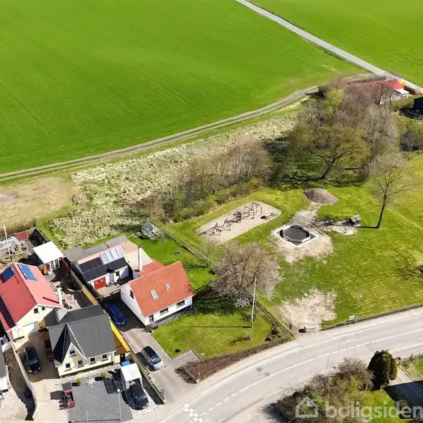 Flere huse med farverige tage ligger langs en vej ved siden af en legeplads. Området er omgivet af grønne marker og træer, hvilket skaber en landlig atmosfære.