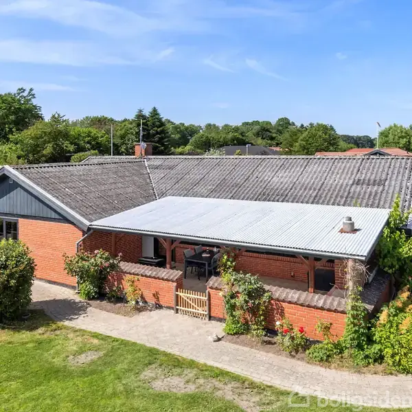En etplans murstenshus med en overdækket terrasse omkranset af buske, beliggende på en grøn, velplejet græsplæne med udsigt til et skovbeklædt område i baggrunden.