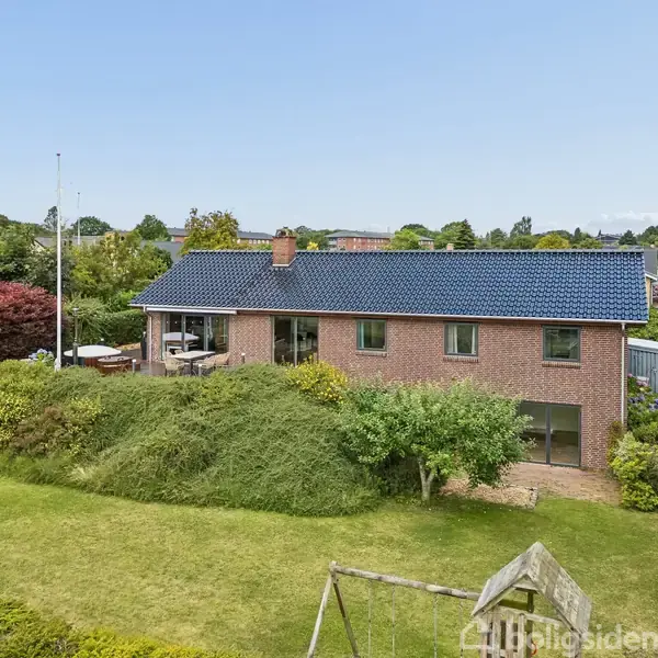 Et rødt murstenshus med blåt tag står i en grøn have med trampolin og gyngestativ. Omkring huset er buske og træer. Horizonterne viser et kvarter med flere huse.