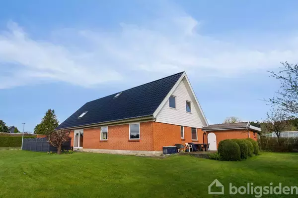 Et rødt murstenshus med mørkt tag står i en grøn have med hække og træer omkring. Huset har flere vinduer og en terrasse med en ståltrappe.