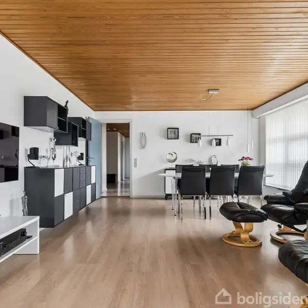 Stue med sort lænestol og fodskammel, sofabord, og spisebord med seks stole. Vægmonteret reol og fladskærms-TV. Træloft og store vinduer med persienner. Klar og minimalistisk indretning.