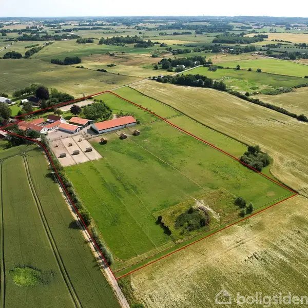 Luftfoto af gård beliggende i det åbne landskab, omgivet af marker under en klar himmel. Gården består af flere bygninger med røde tage og grønne marker afgrænset med rød markering.