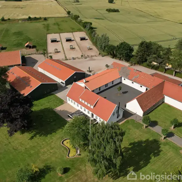 Gårdkompleks med flere rødtagsbygninger danner en gårdsplads. Det omkringliggende landskab består af grønne marker og træer. Nytilplantet haveområde foran hovedbygningen. Landlig kontekst med åbne marker.