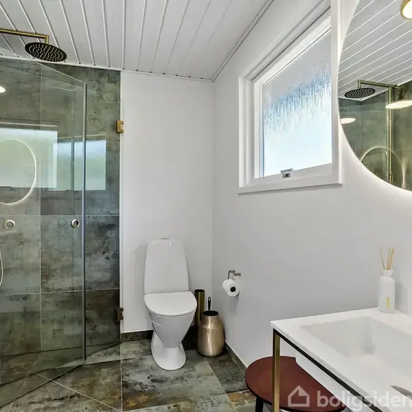 Toilet spejler sig i det glaserede brusekabine hjørne, omgivet af hvide vægge og flisegulv. Håndvask med rund spejl og guldhane i højre side. Vindue giver naturligt lys.