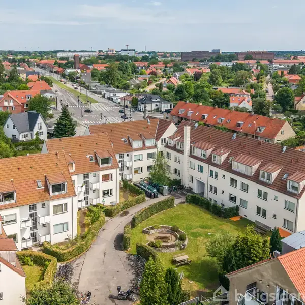 Et lejlighedskompleks med røde tage omkranser en grøn gårdhave. Bygningen er i en by, omgivet af veje, huse og træer, med en landskabelig udsigt i baggrunden.