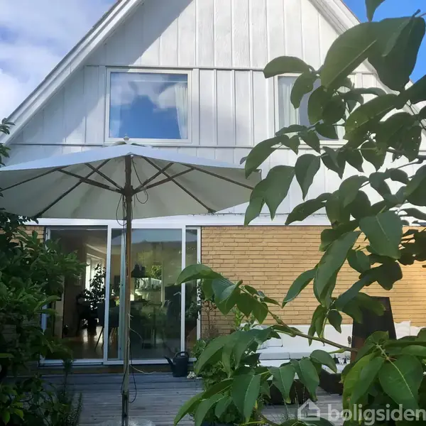 En stor parasol står på en terrasse foran et hus. Huset har hvide og gule murstensdetaljer. Omgivet af grønne planter og træer, med et vindue og skydedør i baggrunden.