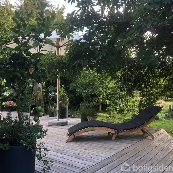 En liggestol er placeret på en træterrasse omringet af frodige grønne planter. En parasol står ved siden af, hvilket skaber en skyggefuld og afslappende atmosfære.