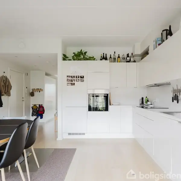 Et moderne køkken med hvide skabe og indbygget ovn. Køkkenbordet har rene linjer, og der er bøger og flasker på overskabene. Et spisebord med sorte stole står til venstre.