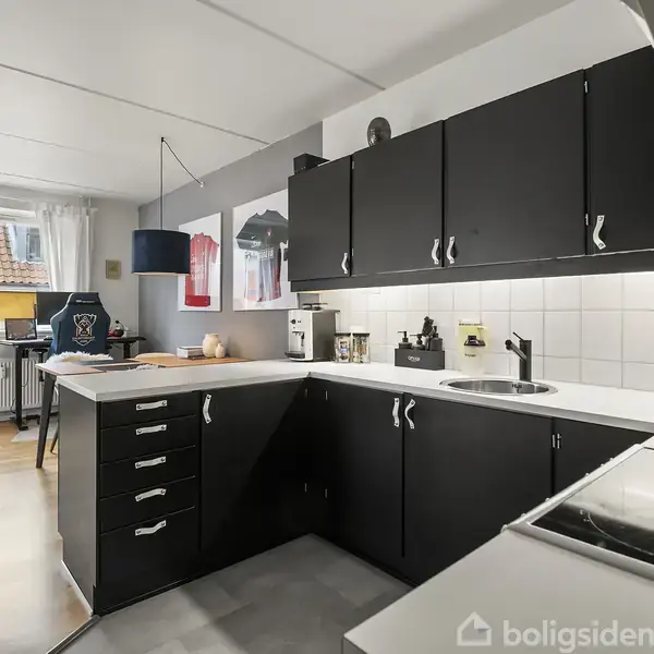 Sort-hvide køkken skabe flankerer vægge, mens en sort bordplade med vask og kogeplader står i fokus. Vindue med hvidt gardin i baggrunden slipper lys ind i rummet.