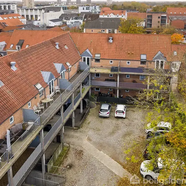 Et toetagers rækkehuskompleks med røde teglstenstage står omkring en gårdsplads med parkerede biler. Omkringliggende bygninger og høstfarvede træer ses i baggrunden.